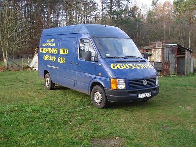 Niebieski van marki Volkswagen z żółtym napisem 'EURO-TRANS BUD' i numerem telefonu 668343638, zaparkowany na trawiastym terenie w pobliżu zardzewiałej szopy